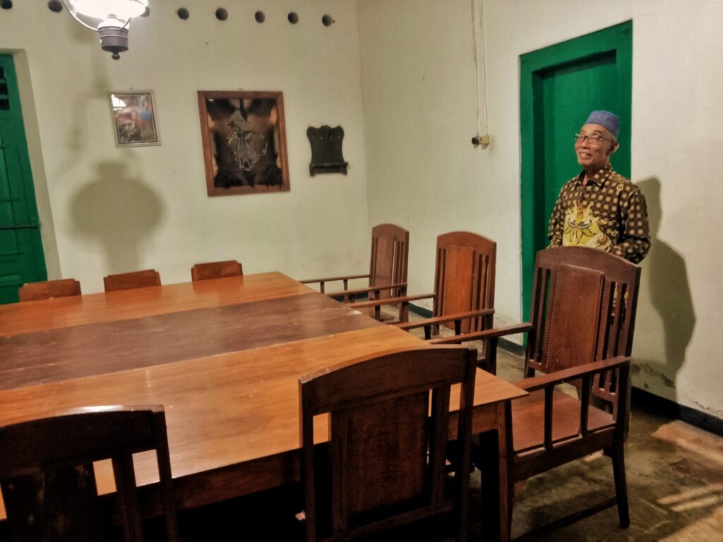 An elderly man in a patterned shirt and blue cap stands beside a green door in a simple room with a long wooden table and chairs nearby